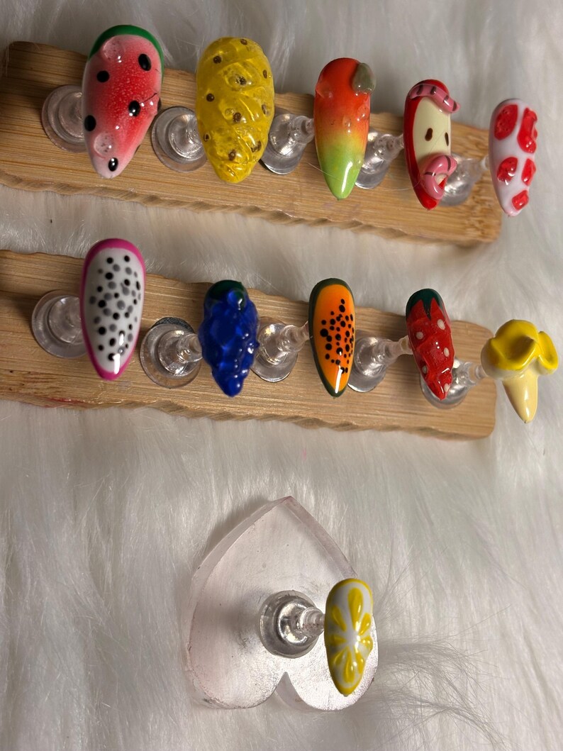 May include: Artificial nails with fruit-themed designs, such as watermelon, pineapple, and dragon fruit. The nails are painted in bright colours and displayed on wooden holders. The nail art features detailed designs and glossy finishes.