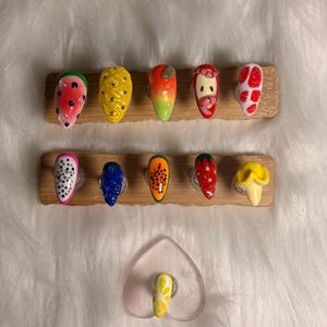 May include: A selection of fruit-themed press-on nails. Designs include watermelon, pineapple, orange, strawberry, and dragon fruit. Each nail is painted with vibrant colours and detailed patterns, mounted on wooden display stands.
