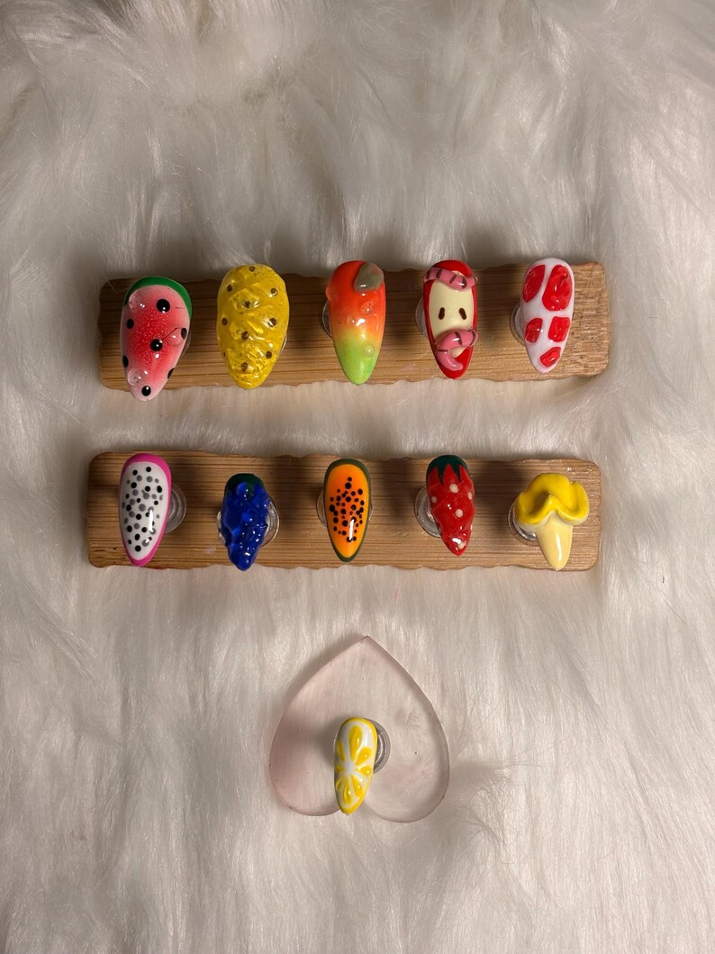 May include: A selection of fruit-themed press-on nails. Designs include watermelon, pineapple, orange, strawberry, and dragon fruit. Each nail is painted with vibrant colours and detailed patterns, mounted on wooden display stands.