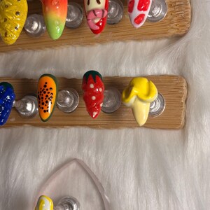 May include: A selection of artificial nails with fruit-themed designs. The nails showcase bright colours and patterns, including a yellow pineapple, an orange and green gradient, a strawberry, and a papaya design. The nails are displayed on wooden holders.