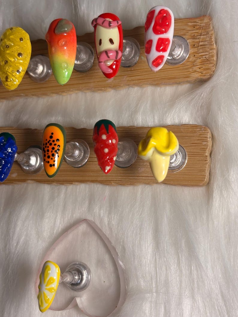 May include: A selection of artificial nails with fruit-themed designs. The nails showcase bright colours and patterns, including a yellow pineapple, an orange and green gradient, a strawberry, and a papaya design. The nails are displayed on wooden holders.