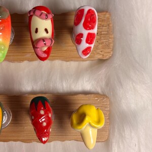 May include: A selection of hand-painted nail art designs in various colours. Designs include a strawberry, a mushroom, and a worm on an apple. The nails are displayed on a wooden board.