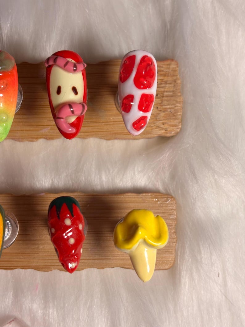 May include: A selection of hand-painted nail art designs in various colours. Designs include a strawberry, a mushroom, and a worm on an apple. The nails are displayed on a wooden board.
