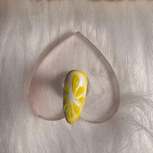 May include: A single acrylic nail featuring a lemon slice design. The nail is painted yellow with white detailing, mimicking a citrus fruit. It rests on a clear, heart-shaped surface against a fluffy white backdrop.