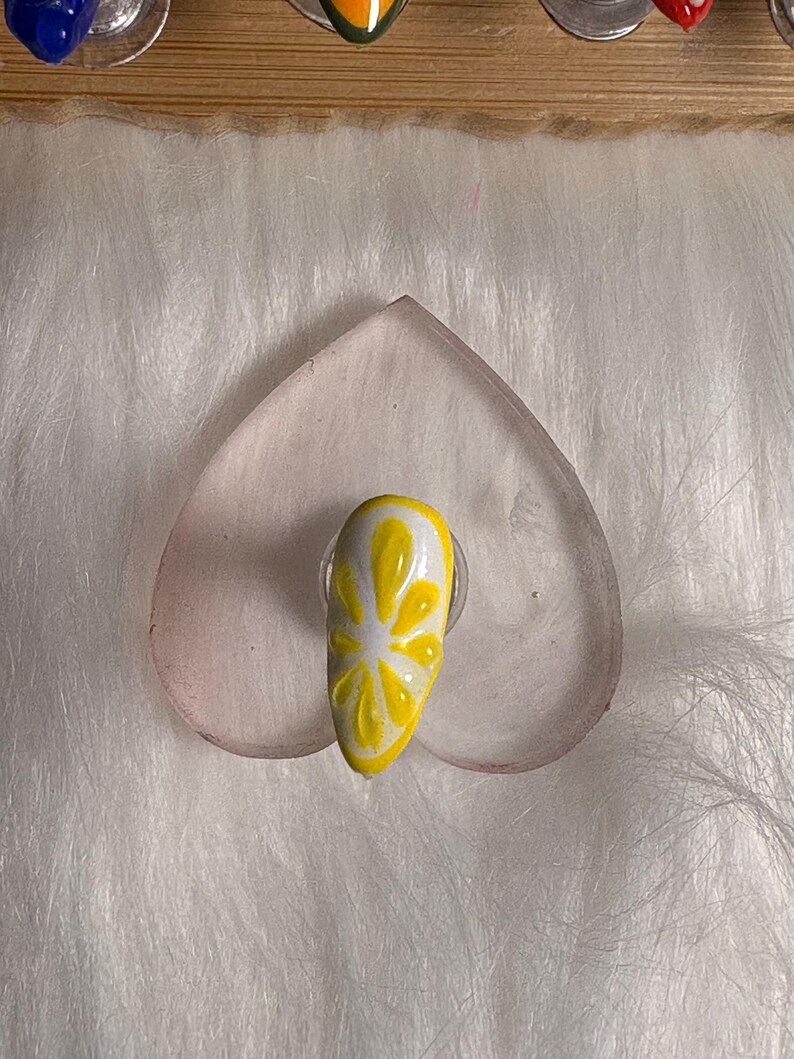 May include: A single acrylic nail featuring a lemon slice design. The nail is painted yellow with white detailing, mimicking a citrus fruit. It rests on a clear, heart-shaped surface against a fluffy white backdrop.