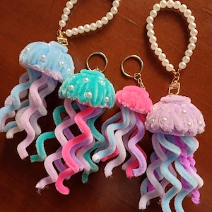 Puede incluir: Cuatro llaveros de medusas coloridas. Cada llavero presenta una cabeza de medusa redonda y afelpada en tonos de azul, turquesa, rosa y morado, adornada con pequeñas perlas blancas. Los tentáculos están hechos de limpiapipas retorcidos y coloridos. Cada llavero tiene un asa de perlas.