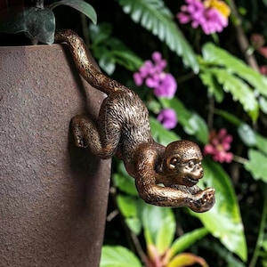 May include: A bronze-colored monkey sculpture clinging to the side of a brown textured pot. The monkey is detailed with a textured fur pattern and is surrounded by green foliage and purple flowers.