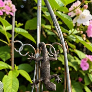 May include: A bronze-colored lizard sculpture is affixed to a decorative metal garden gate. The lizard is detailed with visible claws and textured skin. Pink and white flowers and green foliage are in the background.