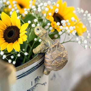 May include: A bronze-colored turtle figurine decorates the rim of a ceramic pot filled with sunflowers and white baby's breath. The turtle has detailed features and appears to be climbing over the edge of the pot, adding a whimsical touch to the floral arrangement.
