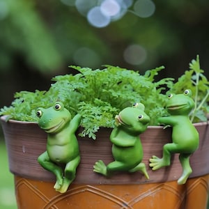 Puede incluir: Tres figuras de ranas verdes adornan el borde de una maceta marrón llena de plantas verdes. Las ranas, con sus caras sonrientes, adoptan diferentes poses, añadiendo un toque caprichoso a la decoración del jardín.