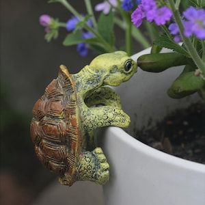 Puede incluir: Una figura decorativa de tortuga en tonos verdes y marrones se aferra al borde de una maceta blanca. El caparazón de la tortuga es marrón, mientras que su cuerpo tiene una apariencia verde texturizada y envejecida. Flores moradas florecen en el fondo.
