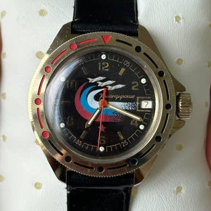 May include: A vintage wristwatch with a black leather band and a gold-toned bezel. The watch face is black with a colorful emblem and white numerals. The watch is displayed on a white, perforated pillow.