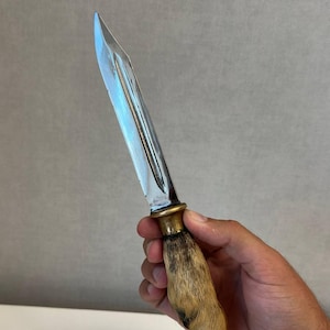 Puede incluir: Un cuchillo de hoja fija con una hoja de acero pulido con un canal central, una guarda de latón y un mango hecho de una pezuña de animal marrón y negra. La hoja es puntiaguda y de doble filo, unida a una virola de tono dorado metálico.