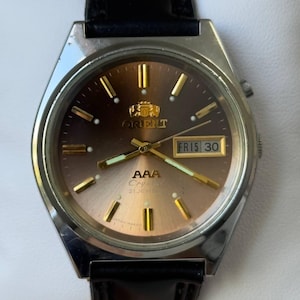 May include: An Orient AAA Crystal 21 Jewels wristwatch with a silver-tone metal case and a black leather strap. The watch features a sunburst brown dial, gold-tone hour markers, gold-tone hands with luminous inserts, and a day-date window at 3 o'clock.