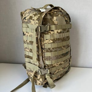 Könnte beinhalten: Ein taktischer Rucksack mit digitalem Tarnmuster in Beige-, Braun- und Grüntönen. Der Rucksack verfügt über mehrere Reihen horizontaler Gurtbänder, seitliche Kompressionsriemen mit Kunststoffschnallen und ein kleines weißes Etikett an der Oberseite.