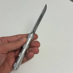 Op de afbeelding: Een zilverkleurig metalen hobbymes met een slank, puntig lemmet en een cilindrische handgreep met een getextureerd, gekarteld patroon. Het gereedschap is volledig in beeld, met een metalen liniaal gedeeltelijk zichtbaar in de hoek.