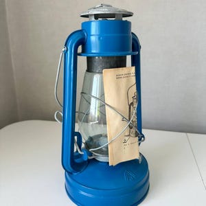 May include: A blue vintage lantern with a clear glass globe and a silver top. The lantern has a handle for carrying and a small paper tag attached. The lantern is designed to hold a fuel source for illumination.