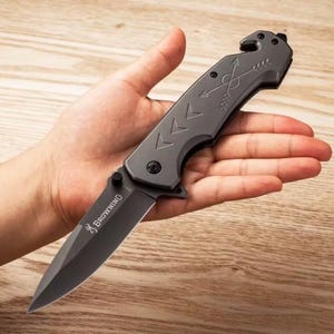 Puede incluir: Una navaja gris con una hoja negra. La navaja tiene un mango con dise&ntilde;os de flechas y galones y la marca "Browning" impresa en la hoja. La navaja se sostiene abierta en la mano de una persona.