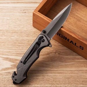 Puede incluir: Una navaja gris con una hoja afilada y un clip para el bolsillo. El mango presenta un dise&ntilde;o de flecha. La navaja est&aacute; sobre una superficie de madera, con una caja de madera en el fondo.