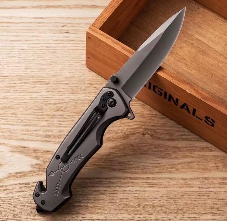 Puede incluir: Una navaja gris con una hoja afilada y un clip para el bolsillo. El mango presenta un dise&ntilde;o de flecha. La navaja est&aacute; sobre una superficie de madera, con una caja de madera en el fondo.