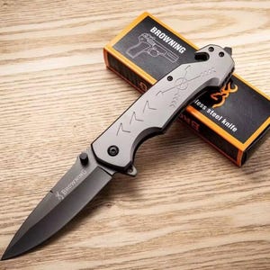 Puede incluir: Una navaja plegable Browning gris y negra con una hoja de acero inoxidable. El cuchillo tiene un mango texturizado con dise&ntilde;os de flechas y chevrones. La caja detr&aacute;s del cuchillo es naranja y negra y presenta el logotipo de Browning y una ilustraci&oacute;n de pistola.