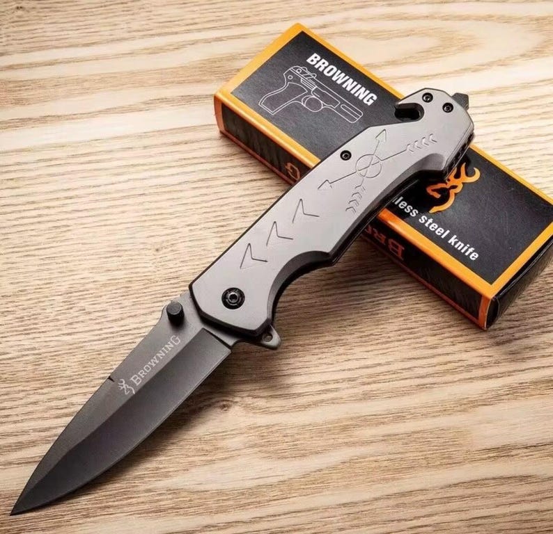 Puede incluir: Una navaja plegable Browning gris y negra con una hoja de acero inoxidable. El cuchillo tiene un mango texturizado con dise&ntilde;os de flechas y chevrones. La caja detr&aacute;s del cuchillo es naranja y negra y presenta el logotipo de Browning y una ilustraci&oacute;n de pistola.