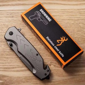 Puede incluir: Una navaja plegable de acero inoxidable gris con dise&ntilde;os de flechas y br&uacute;jula, junto a su caja negra y naranja. La caja dice "BROWNING Cuchillo de acero inoxidable" con una ilustraci&oacute;n de pistola y un logotipo de ciervo.