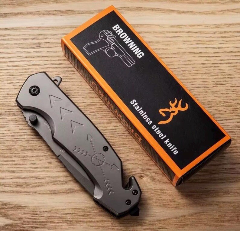 Puede incluir: Una navaja plegable de acero inoxidable gris con dise&ntilde;os de flechas y br&uacute;jula, junto a su caja negra y naranja. La caja dice "BROWNING Cuchillo de acero inoxidable" con una ilustraci&oacute;n de pistola y un logotipo de ciervo.