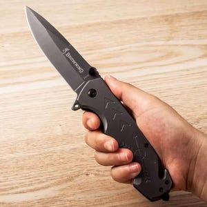 Puede incluir: Una navaja plegable gris oscuro con una hoja puntiaguda y un mango texturizado. La navaja tiene la palabra "Browning" impresa en la hoja. El mango presenta dise&ntilde;os en forma de flecha y un peque&ntilde;o gancho en la base.