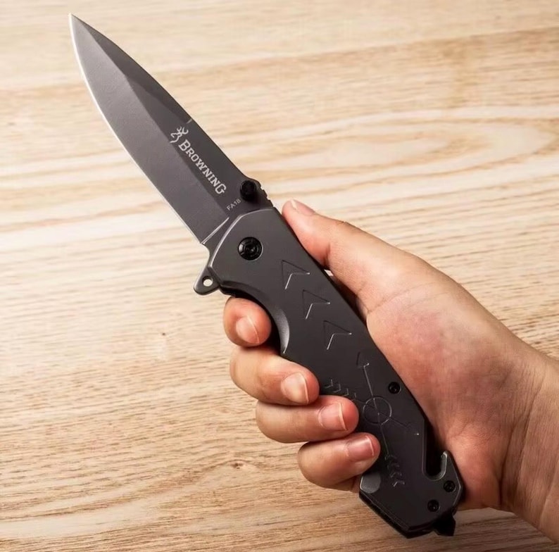Puede incluir: Una navaja plegable gris oscuro con una hoja puntiaguda y un mango texturizado. La navaja tiene la palabra "Browning" impresa en la hoja. El mango presenta dise&ntilde;os en forma de flecha y un peque&ntilde;o gancho en la base.