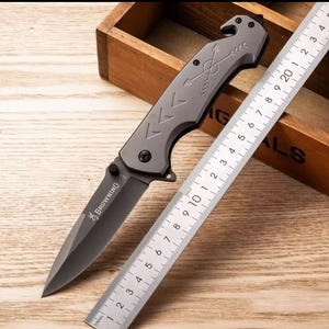 Puede incluir: Una navaja plegable gris con una hoja negra y la palabra "BROWNING" impresa. La navaja tiene un dise&ntilde;o de flecha en el mango y est&aacute; junto a una regla marcada en cent&iacute;metros. La navaja est&aacute; sobre una superficie de madera.