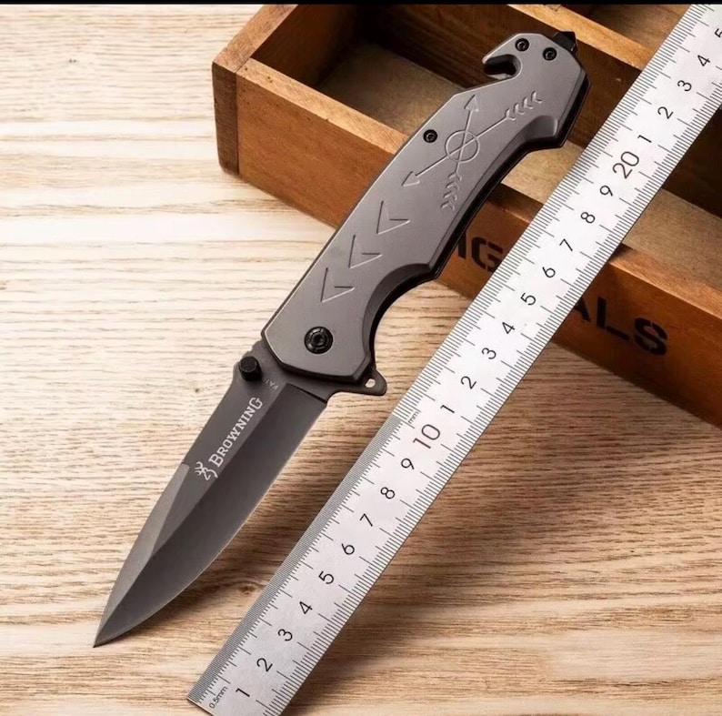 Puede incluir: Una navaja plegable gris con una hoja negra y la palabra "BROWNING" impresa. La navaja tiene un dise&ntilde;o de flecha en el mango y est&aacute; junto a una regla marcada en cent&iacute;metros. La navaja est&aacute; sobre una superficie de madera.