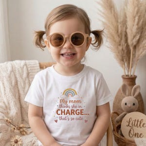 Puede incluir: Una niña pequeña con una camiseta blanca con el texto "My mom thinks she in CHARGE... that's so cute" y un arcoíris. La niña lleva gafas de sol redondas de color marrón claro y sonríe. El fondo incluye una silla de mimbre, hierba de la pampa y un letrero de madera.