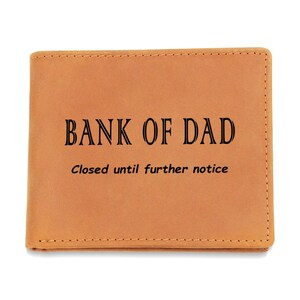 Puede incluir: Una cartera de cuero marrón claro con el texto "BANK OF DAD" en negro y la frase "Closed until further notice" debajo. La cartera es rectangular y tiene bordes cosidos.