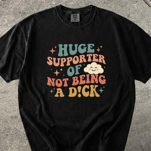 May include: Black t-shirt featuring the phrase "HUGE SUPPORTER OF NOT BEING A D!CK" in a retro font. The shirt has a faded appearance. A cat wearing sunglasses is in the upper right corner.