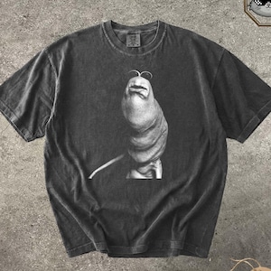May include: Dark gray t-shirt featuring a black and white illustration of a stylized, abstract figure. The word "Pepper" is printed in cursive at the bottom left. The shirt has a vintage, worn look.
