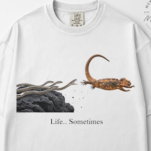Op de afbeelding: Een wit T-shirt met korte mouwen en een grafische print van een hagedis die wegspringt van een donkere rots waar verschillende slangen uit tevoorschijn komen. Onder de afbeelding staat de tekst "Life.. Sometimes". Ronde hals en losse pasvorm.