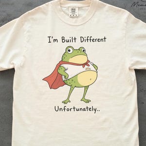 May include: An ivory short-sleeved t-shirt featuring a graphic of a green frog wearing a white shirt and a red cape. The text above the frog reads I'm Built Different and the text below reads Unfortunately.. The shirt has a crew neckline.