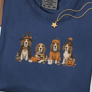 May include: A true navy blue t-shirt featuring a graphic of four beagle dogs dressed in Halloween costumes, including a witch, a pumpkin, and winter accessories. The shirt has a Comfort Colors label in the neck and a gold star pendant necklace.