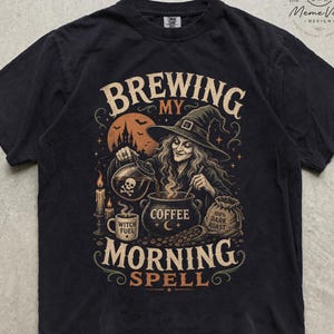 Può includere: Una t-shirt nera con una stampa grafica di una strega che mescola un calderone con la scritta COFFEE. Il testo recita BREWING MY MORNING SPELL, con una tazza con la scritta WITCH FUEL e un sacchetto con 100% DARK ROAST. Il design include un castello.
