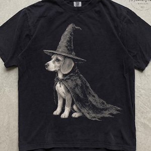 Może przedstawiać: Czarny t-shirt z krótkim rękawem z grafiką przedstawiającą psa rasy beagle w spiczastym kapeluszu czarownicy i ciemnej pelerynie. Wzór znajduje się na środku klatki piersiowej, a koszulka ma okrągły dekolt i standardowy krój.