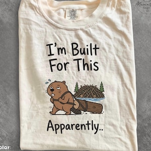 May include: An ivory t-shirt featuring a graphic of a brown beaver pulling a large log. The text reads "I'm Built For This Apparently.." above and below the beaver illustration. The shirt has a crew neckline and short sleeves, with a Comfort Colors label.