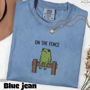May include: A blue jean coloured t-shirt featuring a graphic of a green frog sitting on a wooden fence with the text ON THE FENCE printed above it. The shirt has a Comfort Colors label at the neckline and is paired with a gold star pendant necklace.