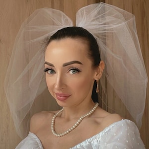 Op de afbeelding: Een vrouw die een witte off-shoulder jurk draagt met een witte tule sluier. Ze draagt een parelketting en oorbellen.