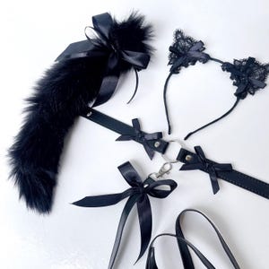 May include: A black cat-themed accessory set featuring a faux fur tail with a satin bow, a headband with lace cat ears, and a collar with a leash. The set includes satin bows and a silver-colored clasp.