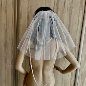 Op de afbeelding: Een witte tule bruids sluier met een delicate glans. De sluier is kort en heeft een enkele laag. Het wordt gedragen op een mannequin met een parelketting.