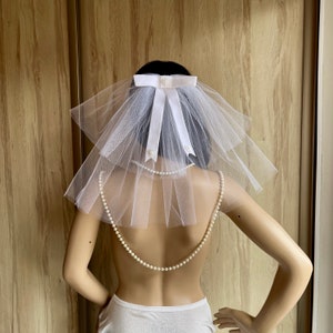 May include: White tulle bridal veil with a white satin bow and pearl accents. The veil is worn on a mannequin with a pearl necklace.