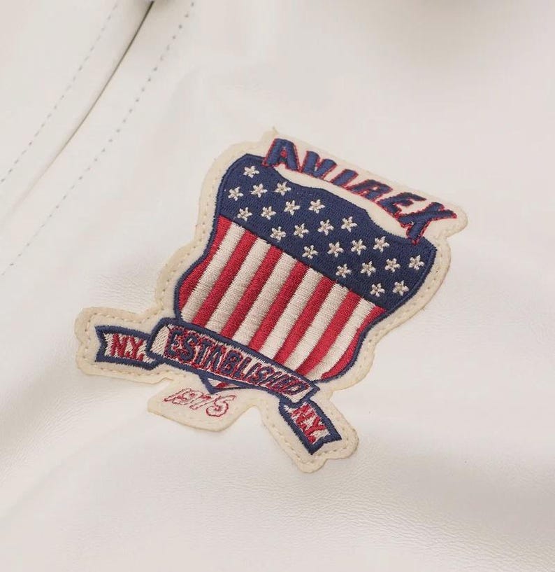 May include: A white leather jacket with a patriotic patch. The patch features the words "AVIREX" and "NY ESTABLISHED 1975" with a design of the American flag. The patch is in red, white, and blue.