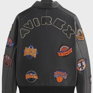 May include: A black leather jacket featuring the word AVIREX in large letters across the upper back. The jacket is decorated with multiple embroidered patches of the New York Knicks basketball team logo in orange, blue, and white colors.