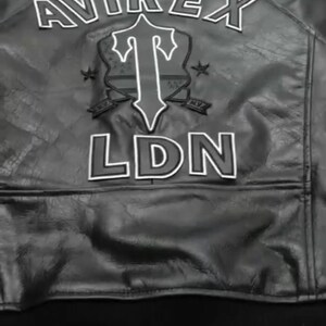 May include: Black leather jacket with white lettering and a shield design. The jacket features the brand name "AVIREX" across the top, with "LDN" at the bottom. A shield graphic is in the center.